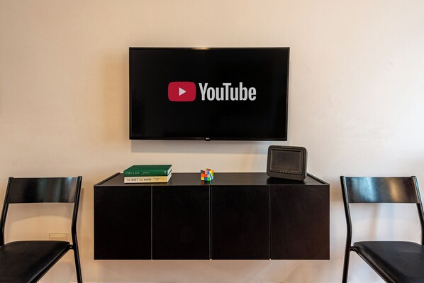 A flat-screen TV mounted on a wall above a black entertainment unit with two black chairs. - Stylish & Charming in Consolação/Higienópolis • Parking • 3rd Guest Free (Sao Paulo)