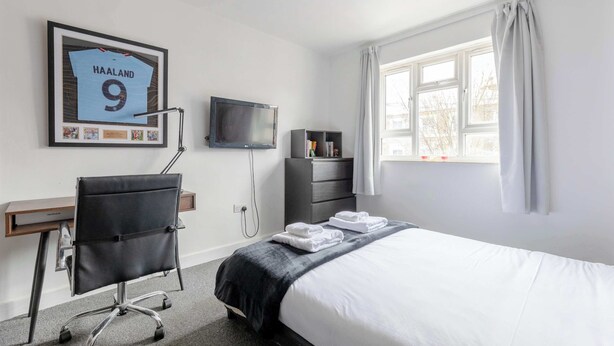 A hotel room with a bed, a desk, a chair, a TV, a window, and a framed jersey on the wall. - Spacious & Central | 1 Bed | WiFi | Regent's Canal (London)