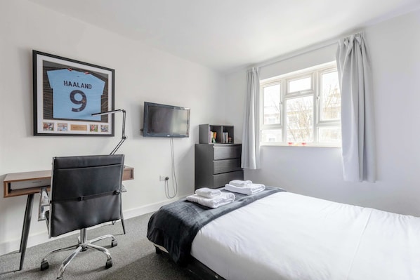 A hotel room with a bed, a desk, a chair, a TV, a window, and a framed jersey on the wall.