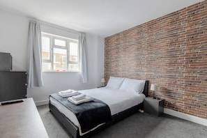 Een hotelkamer met een bakstenen muur, een bed met witte lakens, een bureau met stoel en een raam met gordijnen.