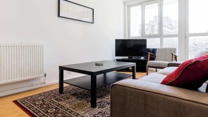 A modern living room with a black coffee table, a grey sofa with a red pillow, a TV, and a large window with a view of buildings.