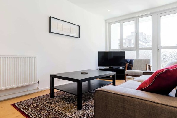 A modern living room with a black coffee table, a grey sofa with a red pillow, a TV, and a large window with a view of buildings.