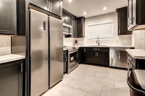 A modern kitchen with dark cabinetry, stainless steel appliances, and a window with blinds.