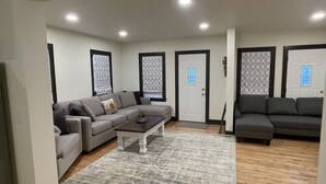 Un salon avec un canapé d’angle gris, une table basse et un tapis gris. Il y a deux portes dotées de panneaux de verre décoratifs et une petite plante accrochée au mur.