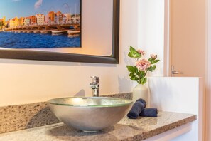 A bathroom with a glass bowl sink, a framed picture of a coastal town, and a vase with flowers.