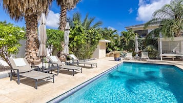 A poolside area with lounge chairs and umbrellas, surrounded by palm trees and a clear blue sky.