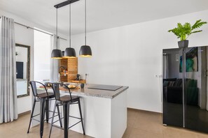 A modern kitchen with a central island, black bar stools, pendant lighting, and a refrigerator with a plant on top.