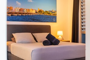 A hotel room with a bed, a nightstand with a lamp, a window with blinds, and a cityscape view on the wall.