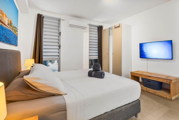 A hotel room with a bed, a TV, a wooden side table, and a window with blinds.