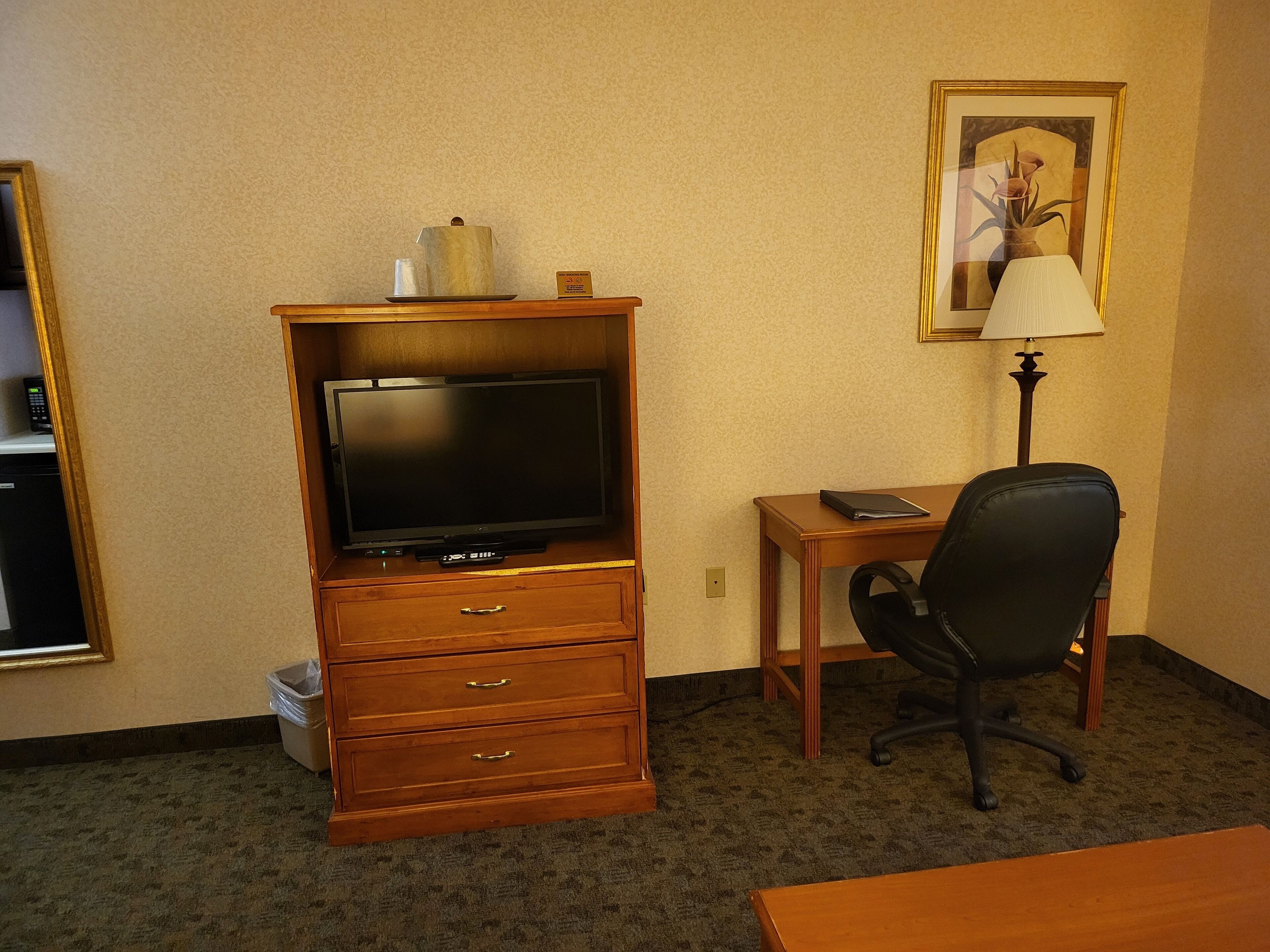 Habitación de hotel con un escritorio de madera, una silla de oficina negra, un televisor sobre un mueble de madera, una lámpara y un cuadro en la pared.