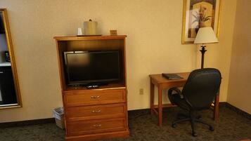 Habitación de hotel con un escritorio de madera, una silla de oficina negra, un televisor sobre un mueble de madera, una lámpara y un cuadro en la pared.