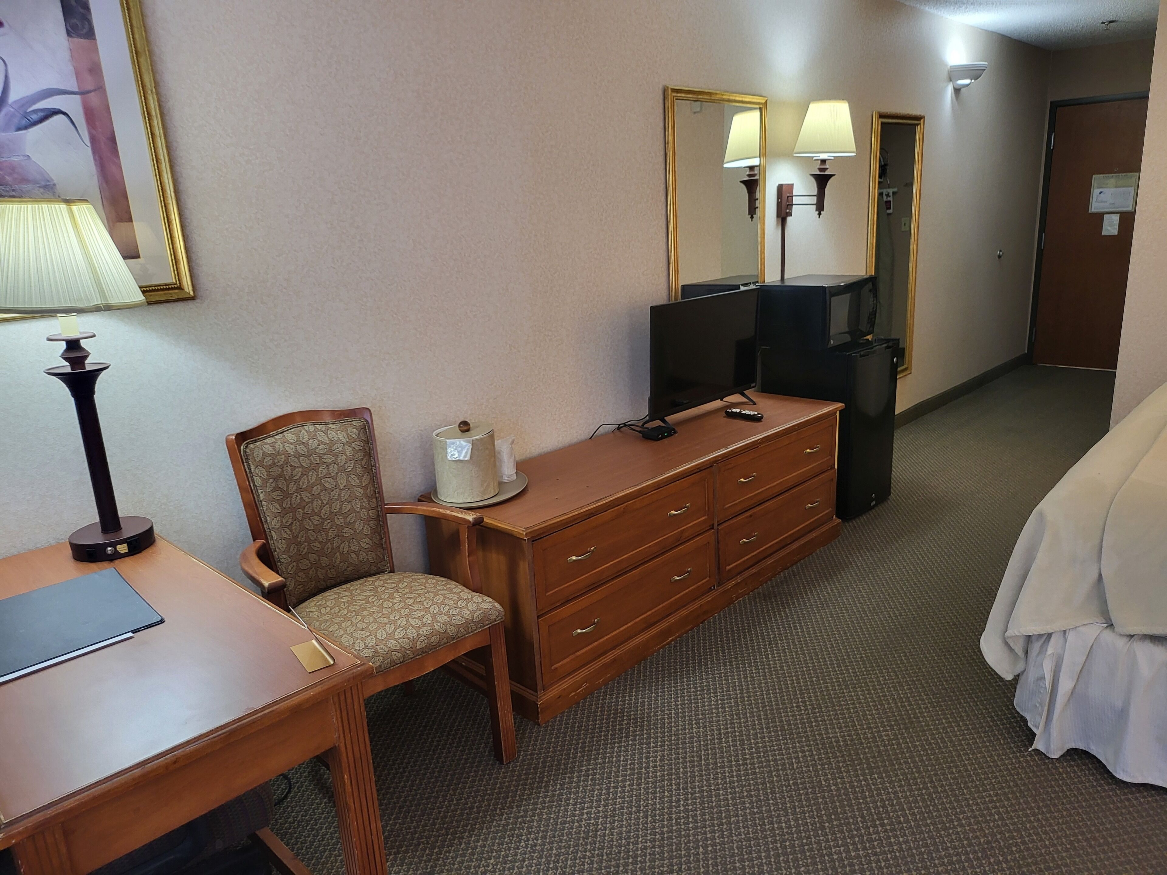 A hotel room with a wooden desk, a chair, a dresser, a television, and a bed.