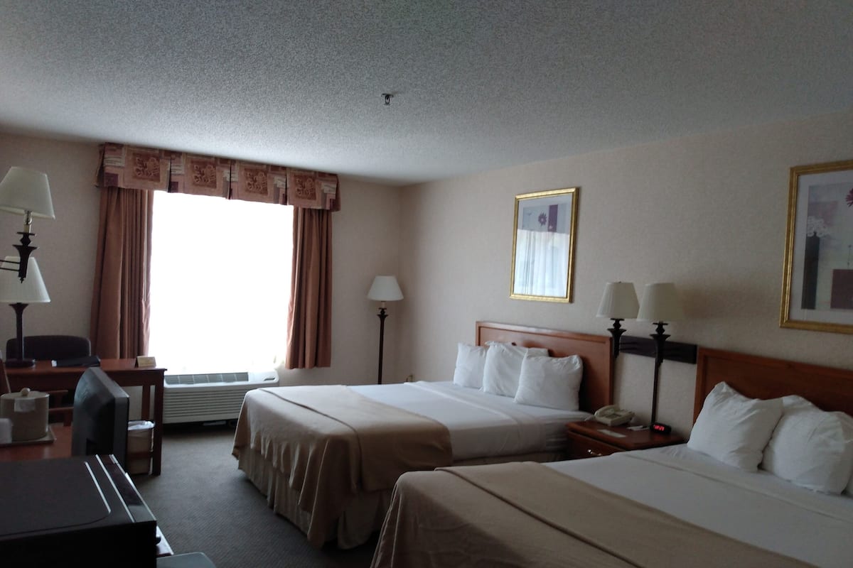 Ein Hotelzimmer mit zwei Betten, einem Schreibtisch, einem Stuhl, einer Lampe und einem Fenster mit Vorhängen.