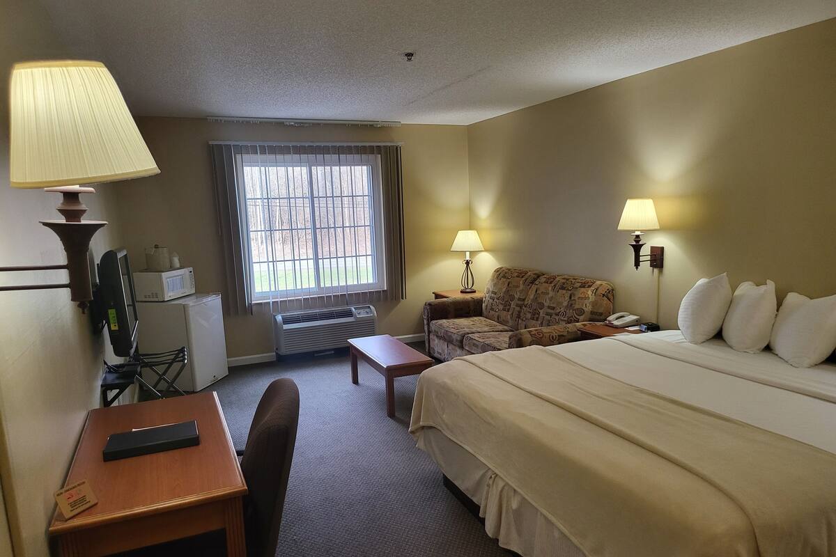 Ein Hotelzimmer mit Bett, Schreibtisch mit Stuhl, Kleintisch, Sofa, Fenster mit Jalousien, Lampe und Mikrowelle.