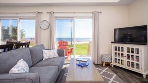 Ein Wohnzimmer mit grauem Sofa, einem Couchtisch, einer TV-Konsole und Blick auf den Strand durch große Glastüren.