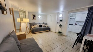 Una habitación moderna y luminosa con un sofá gris, una cama con edredón gris, una mesita con lámpara, y un área de cocina visible a través de una puerta abierta.
