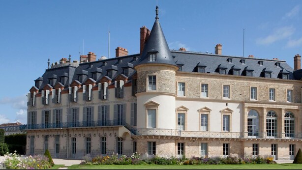 Un edificio grande e histórico con múltiples torres y una fachada simétrica, rodeado de jardines bien cuidados.