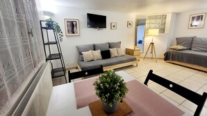 Un salón moderno con un sofá, una mesa de comedor con planta en maceta, un televisor montado en la pared y una ventana con vistas.