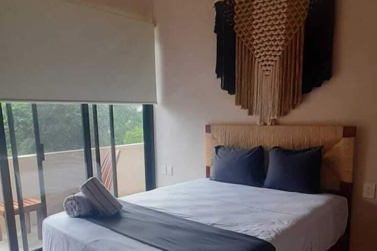 A bedroom with a bed, a ceiling fan, a wall-mounted macrame piece, and a view through a sliding glass door.