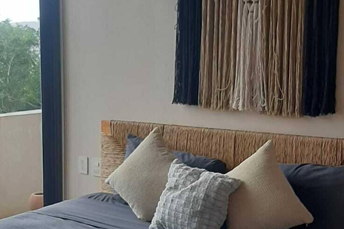 A bedroom with a bed, a headboard with a macrame design, and a view of greenery outside.