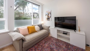 A living room with a sofa, a flat-screen TV, a colorful rug, and a window with a view of trees and buildings. - Lovely Studio with Free Parking in East Melbourne! (East Melbourne)