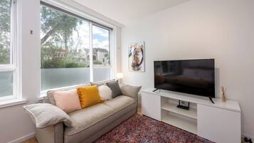 A living room with a sofa, a flat-screen TV, a colorful rug, and a window with a view of trees and buildings.