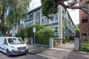 A white van parked in front of a building with a green and beige facade, a staircase, and a sign indicating parking availability.