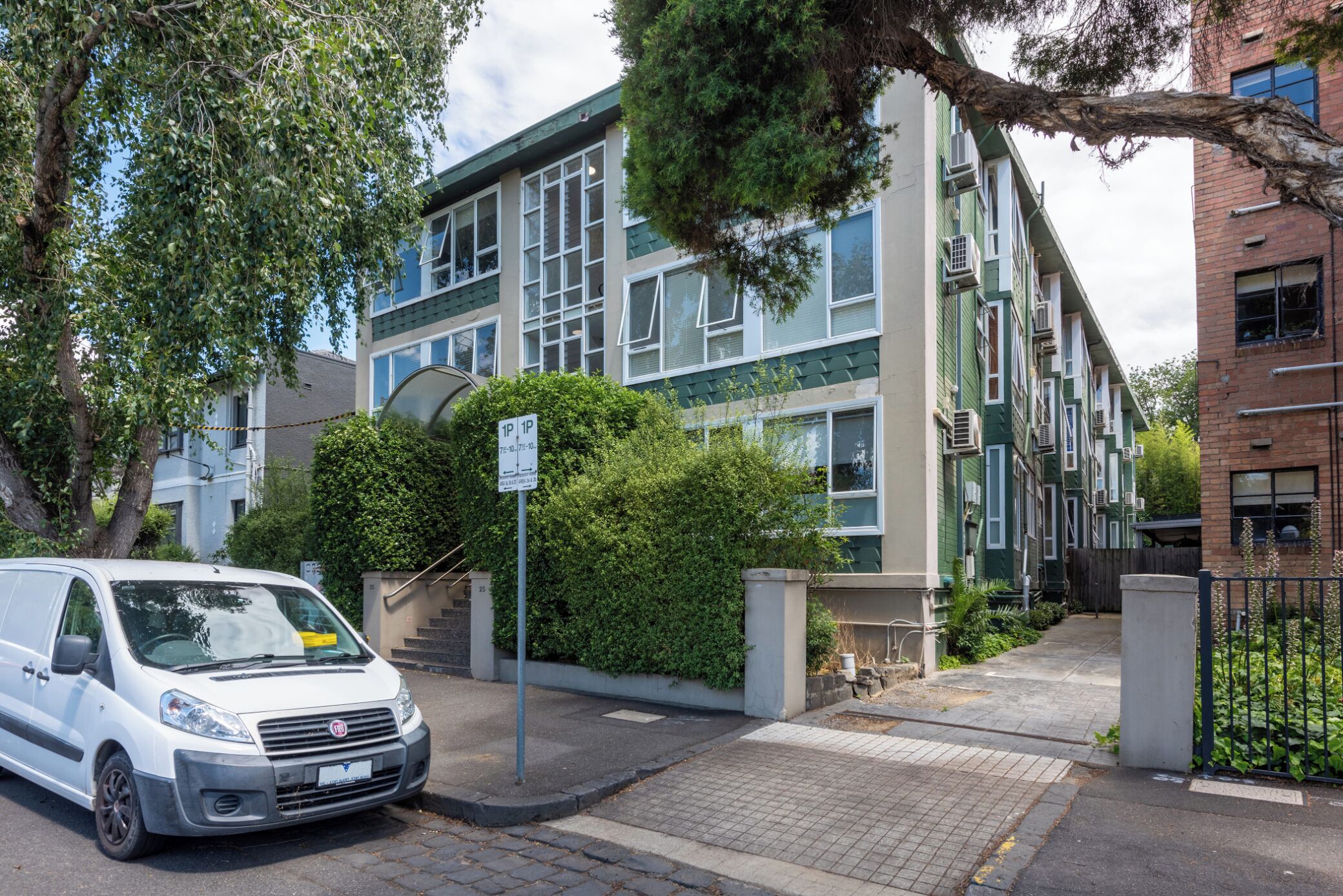 A white van parked in front of a building with a green and beige facade, a staircase, and a sign indicating parking availability.