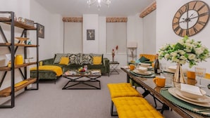 A modern living room with a green sofa, yellow cushions, a coffee table, and a dining area with a table set for two. - Luxurious 1 Bedroom on Iconic Harley Street (Greater London)