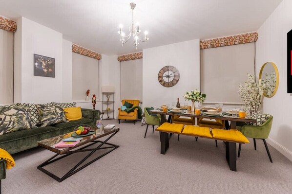 A living room with a green sofa, yellow cushions, a coffee table, and a dining area with a wooden table and chairs.
