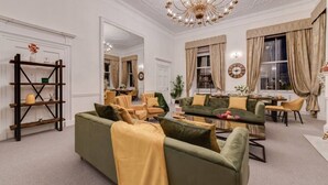 A spacious living room with a large sofa, a coffee table, and a bookshelf. There is a dining area with a table and chairs. The room has a chandelier and large windows with curtains. - Luxury London 3-Bedroom Duplex with Pool Table (Greater London)