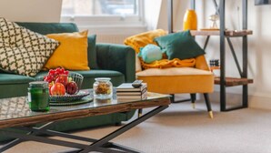 A living room with a green sofa, a yellow armchair, and a coffee table with a fruit bowl, books, and a glass jar.
