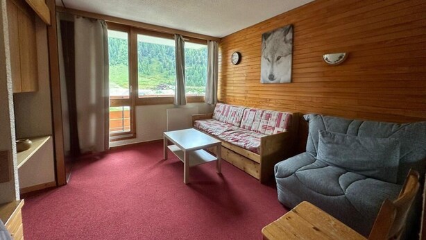 A cozy room with a wooden wall, a sofa, a table, and a window with a view of greenery.