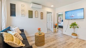 Ein Wohnzimmer mit einem dunkelgrauen Sofa, einem runden Couchtisch mit Blumen und einer Vase, einem flachbildfernseher an der Wand und einem Topf mit Pflanze.