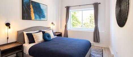 A bedroom with a large bed, a nightstand, a wall-mounted artwork, a window with curtains, and a textured wall decoration.