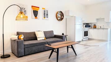 Un salon moderne avec un canapé gris, une table basse en bois et un lampadaire. On y trouve également un coin cuisine avec des meubles blancs et des appareils électroménagers.