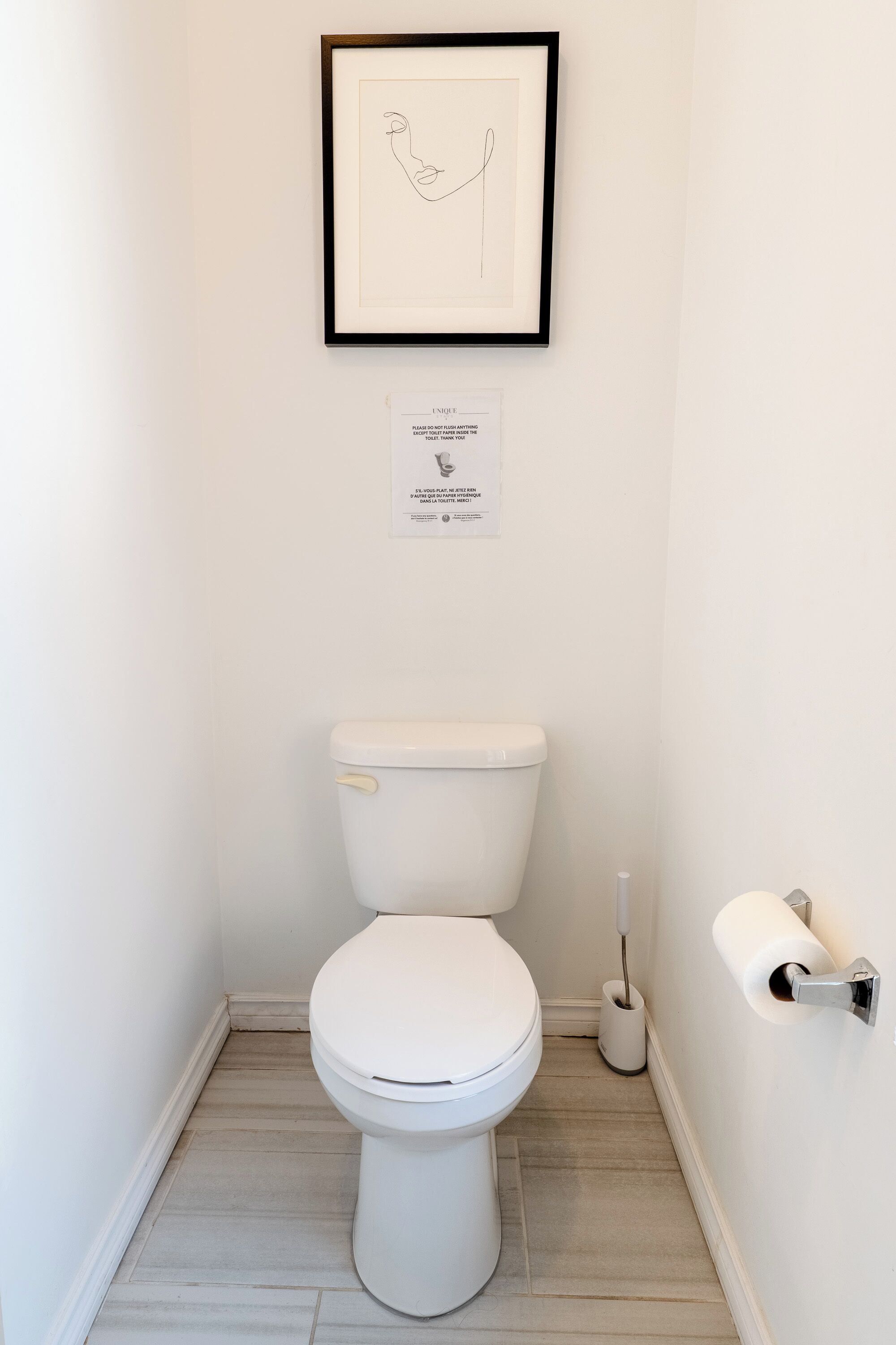 Ein kleines, sauberes Badezimmer mit einer weißen Toilette, einem gerahmten Kunstwerk darüber und einem Schild an der Wand.