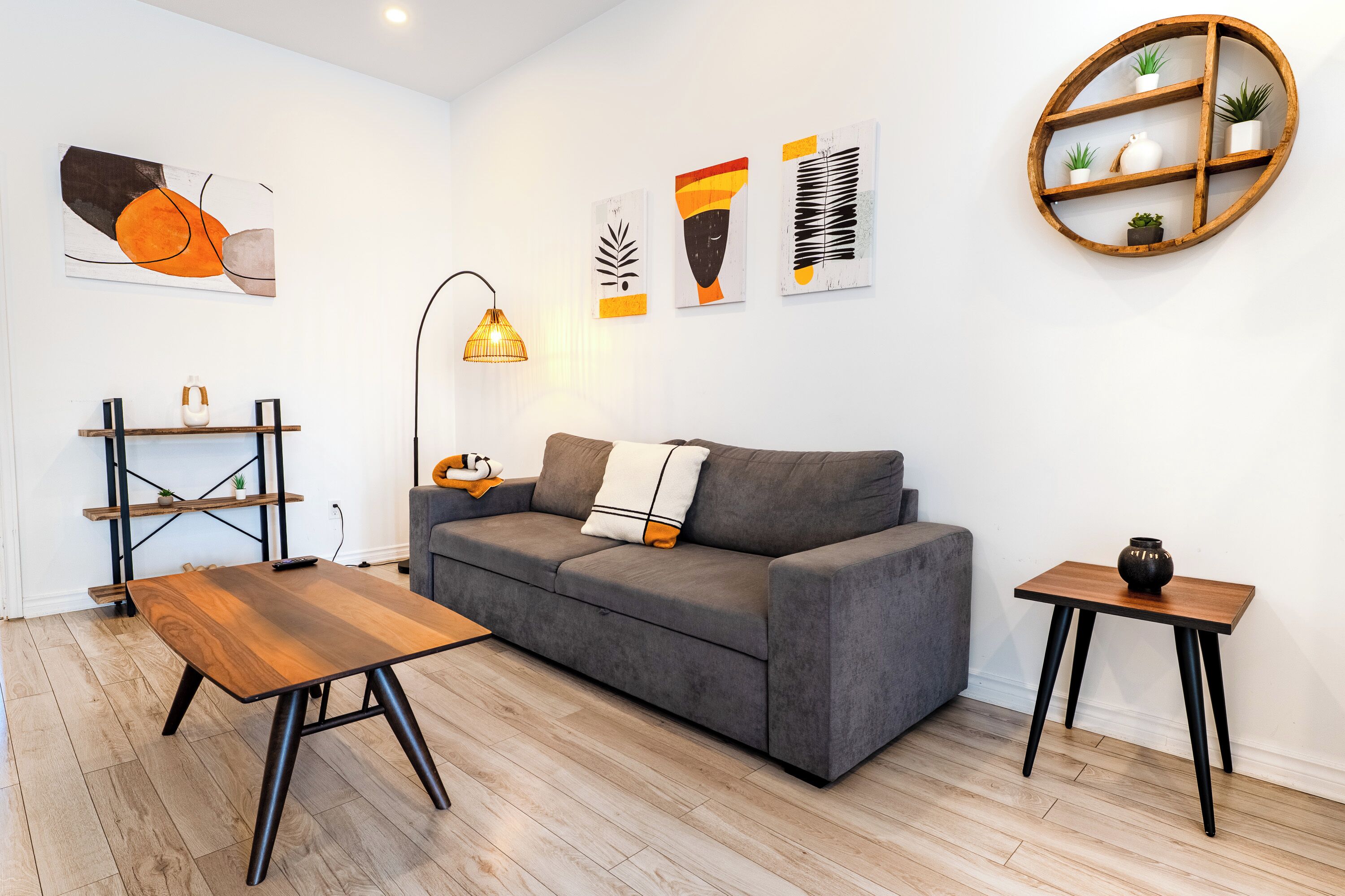 Ein modernes Wohnzimmer mit grauem Sofa, Holztisch und einem kleinen Beistelltisch. An der Wand sind abstrakte Kunstwerke und ein Holzregal mit Pflanzen zu sehen.