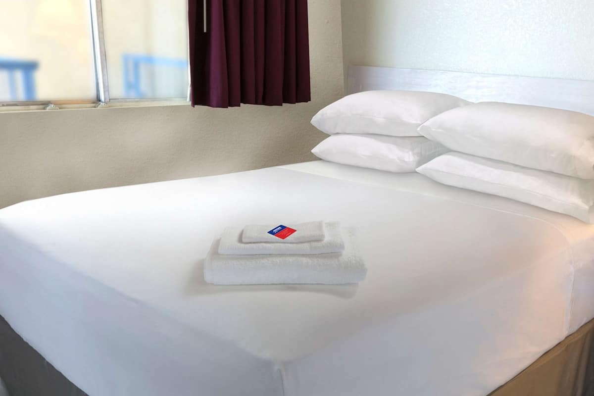 Una cama bien hecha con sábanas y almohadones blancos, una toalla doblada sobre la cama, y una ventana con cortinas.