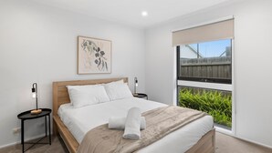Un dormitorio con cama de madera, ropa de cama blanca, una mesita de noche con lámpara, una obra de arte botánica enmarcada en la pared y un ventanal con vistas a la vegetación.