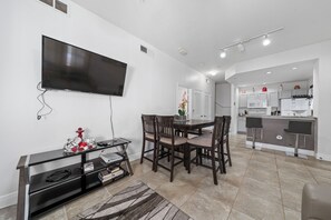 Een moderne woonkamer met een flatscreen televisie, een eethoek met stoelen, een keuken met een bar, en een groot raam.