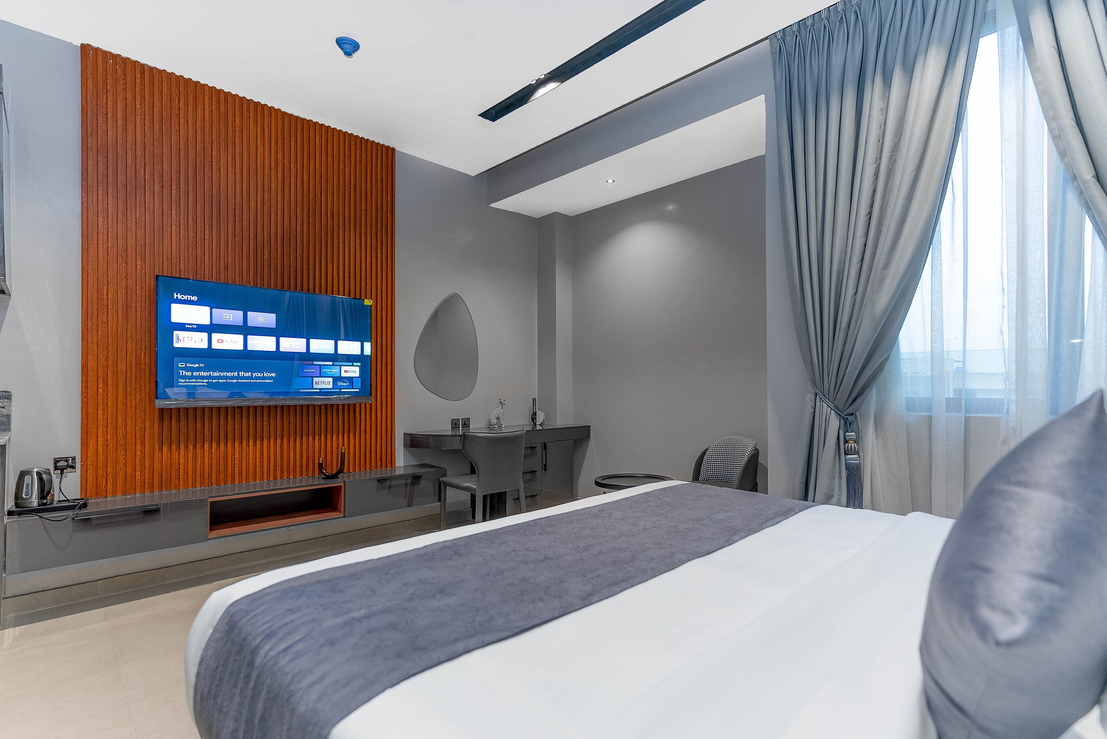 A modern hotel room with a large bed, a flat-screen TV mounted on a wooden panel, a desk with a chair, and a view of the outdoors through sheer curtains.