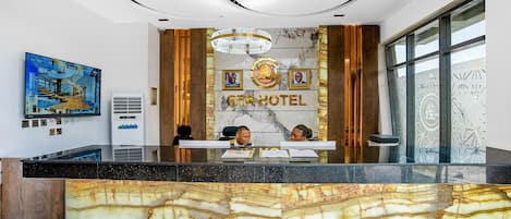 A hotel reception desk with marble countertops, a chandelier, framed photos, and a large window with geometric patterns.
