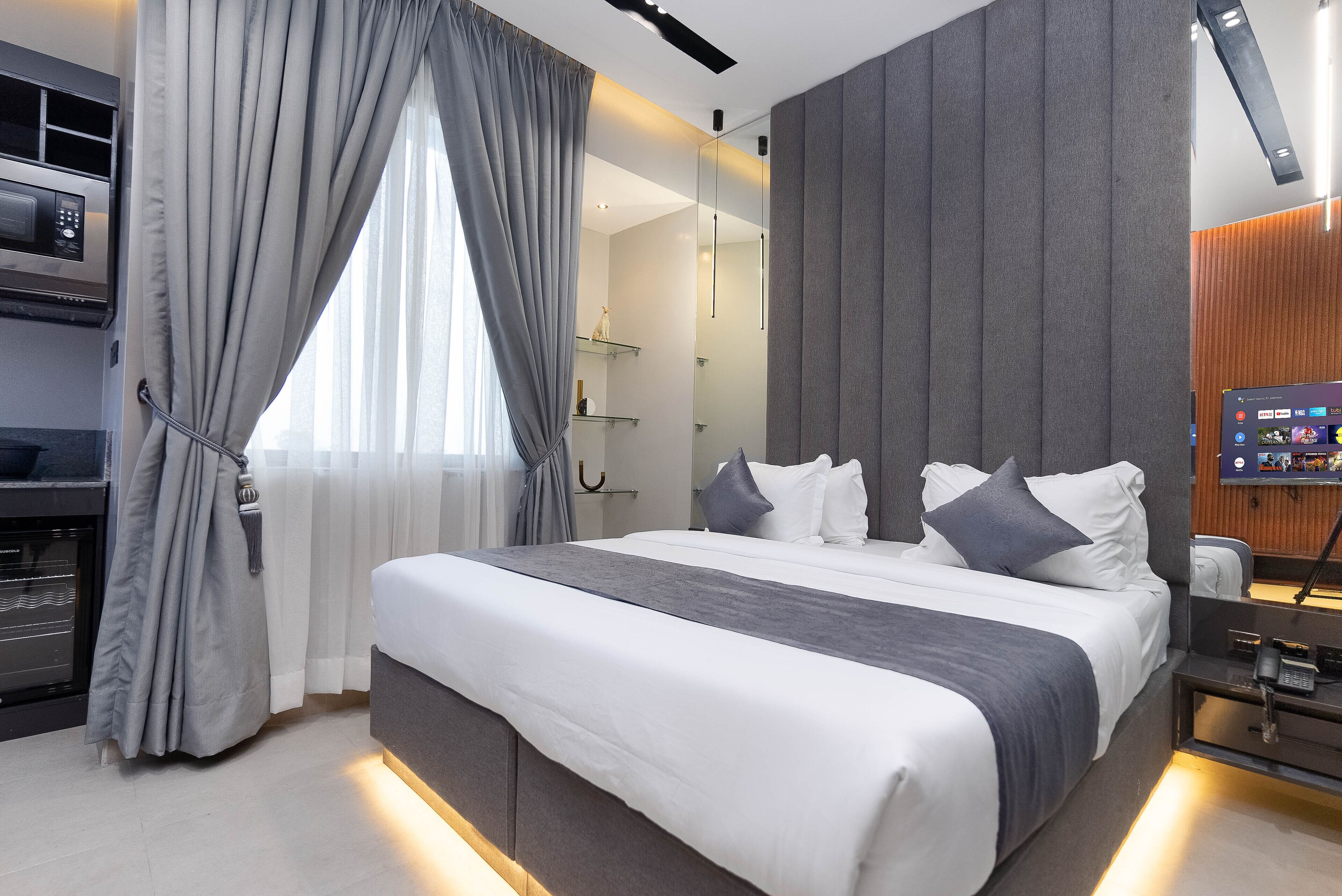 Une chambre d’hôtel moderne avec un grand lit, des rideaux gris, une table de chevet avec une lampe et un téléviseur fixé au mur.