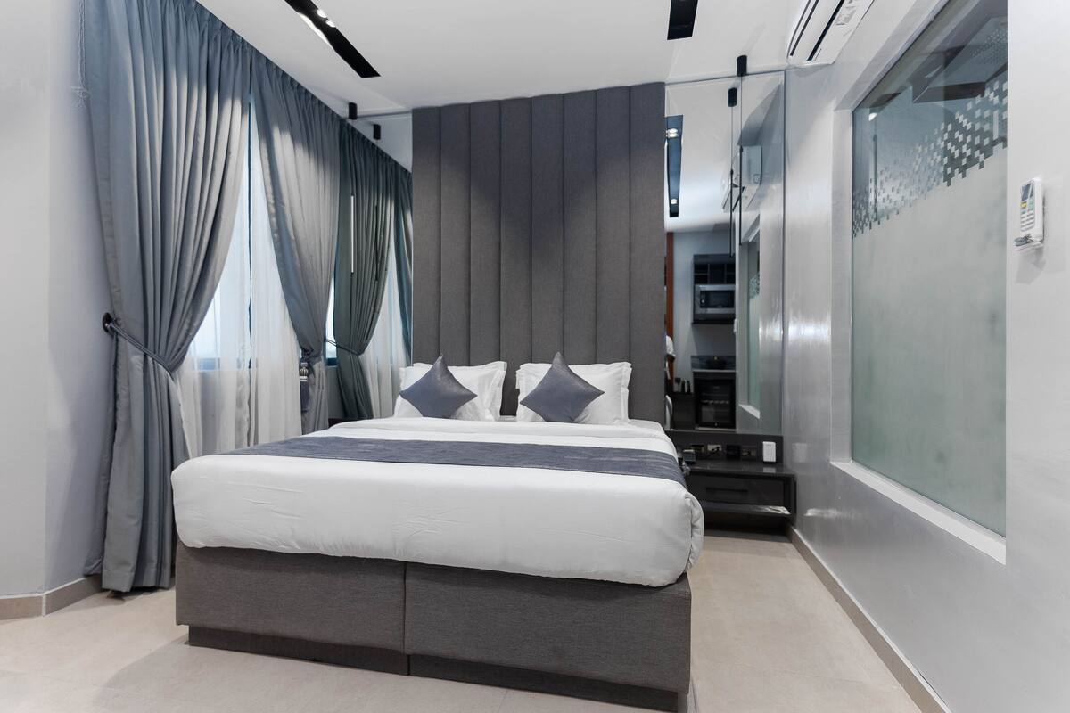 A modern bedroom with a large bed, grey curtains, and a glass partition on one side.