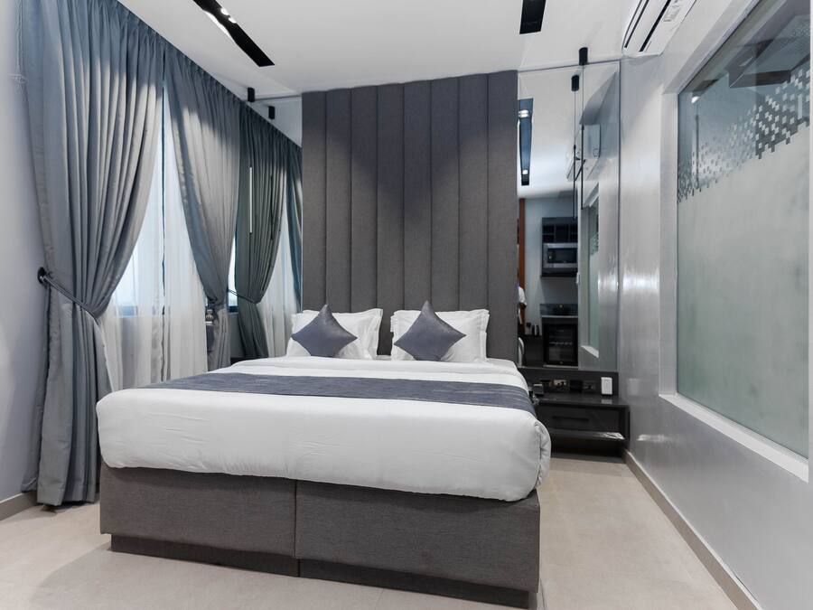 A modern bedroom with a large bed, grey curtains, and a glass partition on one side.