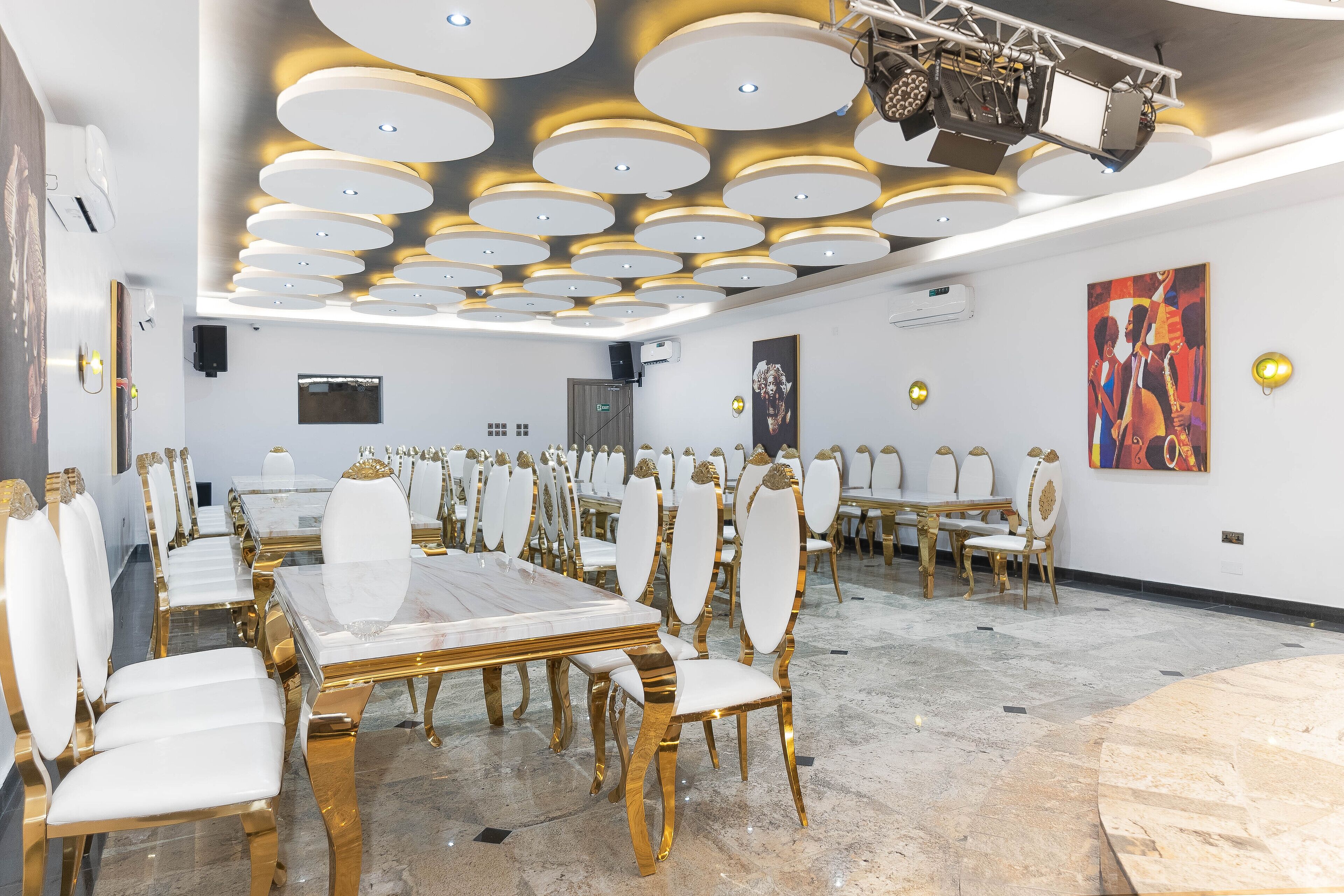 Une pièce spacieuse avec une longue table et des chaises, un plafond orné de luminaires circulaires et un grand tableau accroché au mur.