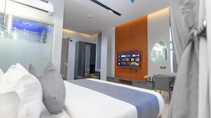 Quarto de hotel moderno com cama, televisão, escrivaninha e casa de banho visível através de uma porta de vidro.