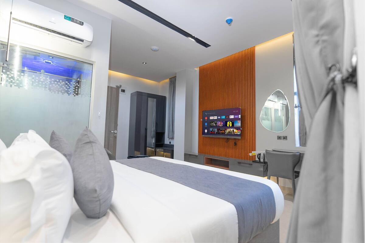 A modern hotel room with a bed, a TV, a desk, and a bathroom visible through a glass door.