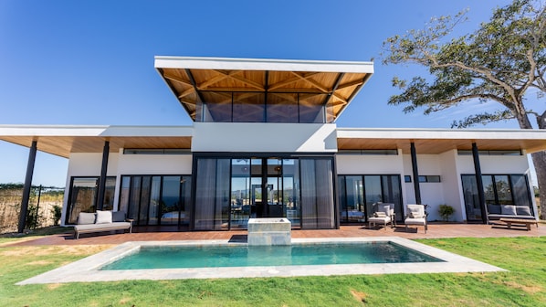 Uma casa moderna de dois andares com fachada em vidro, piscina e área de estar externa.